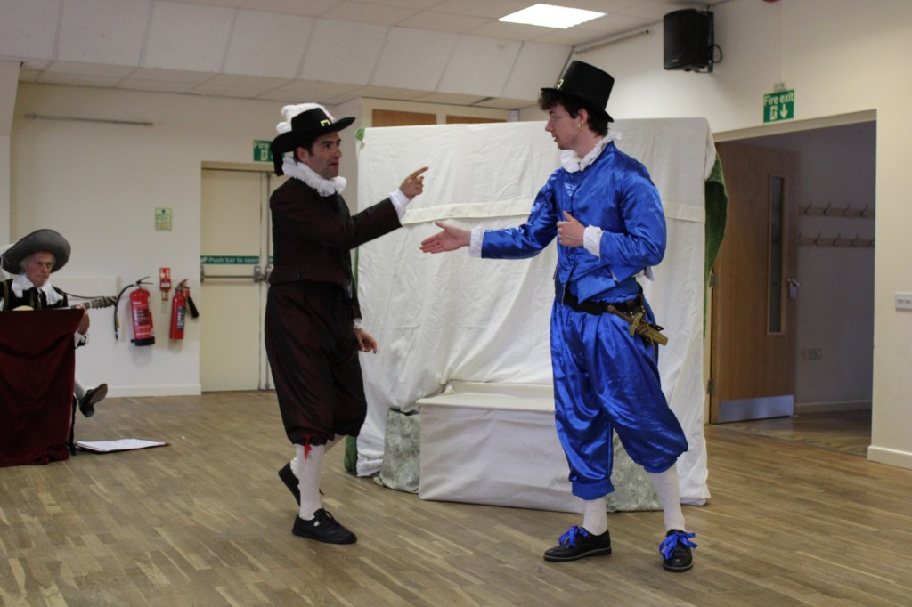 Two men in early modern costume. One points at the other while the other reaches out to shake the first's hand