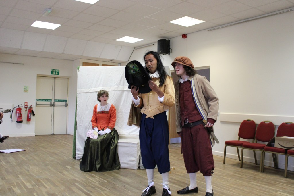 Two men looking inside a hat, while a woman sits in the background