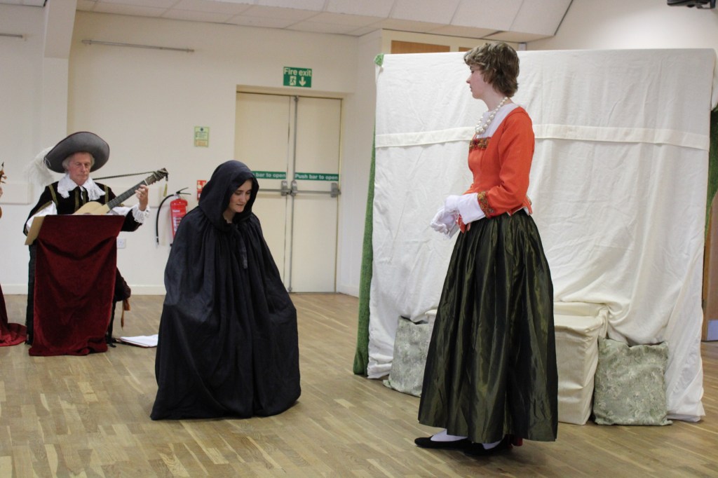 A woman in a cloak curtseys to another woman