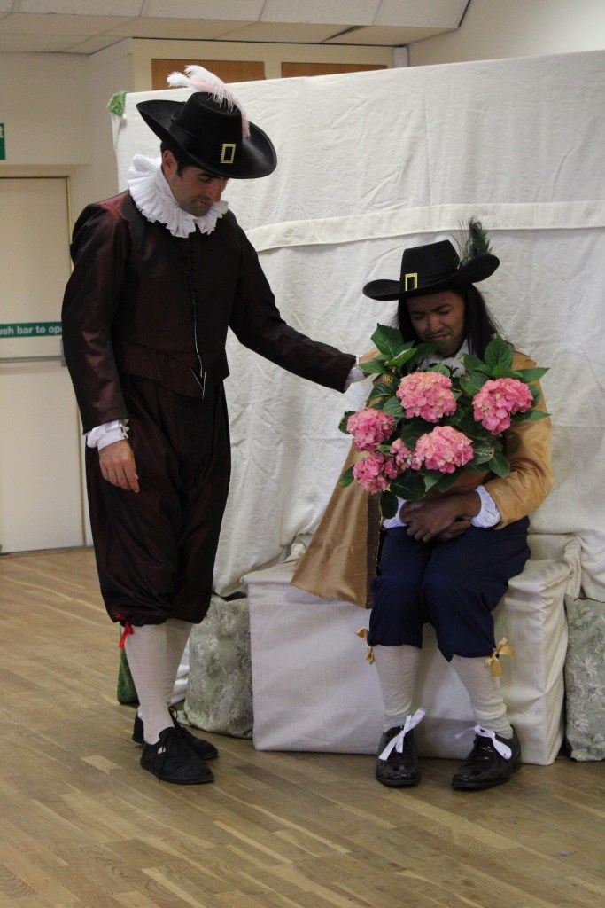 A man stand with his hand on the shoulder of another man who is sitting. The seated man is holding a plant.